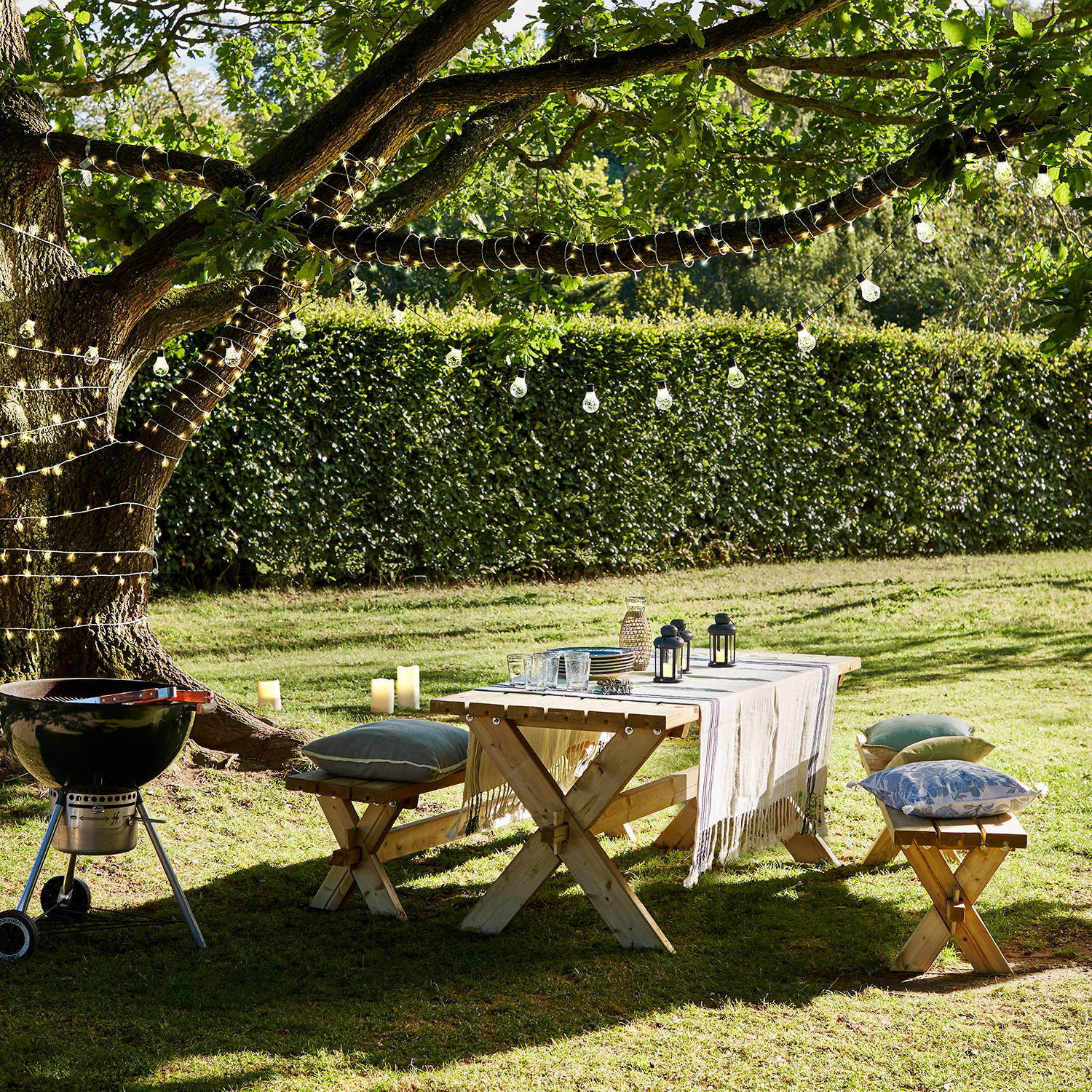 An outdoor summer BBQ setting with fairy lights wrapped around a tree and festoon lights hanging above an outdoor picnic table.