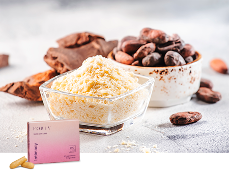 A clear glass bowl of grated cacao butter with a rustic and creamy texture, alongside whole cacao beans and raw chocolate pieces, all set on a light surface with a hint of cocoa powder sprinkled around.