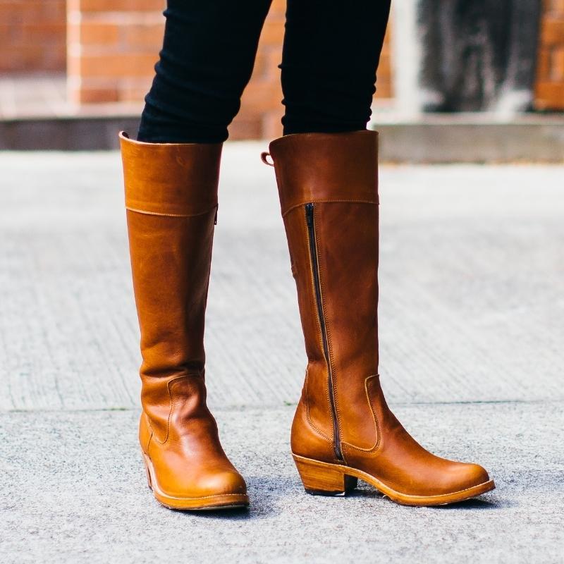 Woman wearing tan leather Condesa wide calf riding boots featuring a full-length side zipper, standing on an urban concrete walkway.