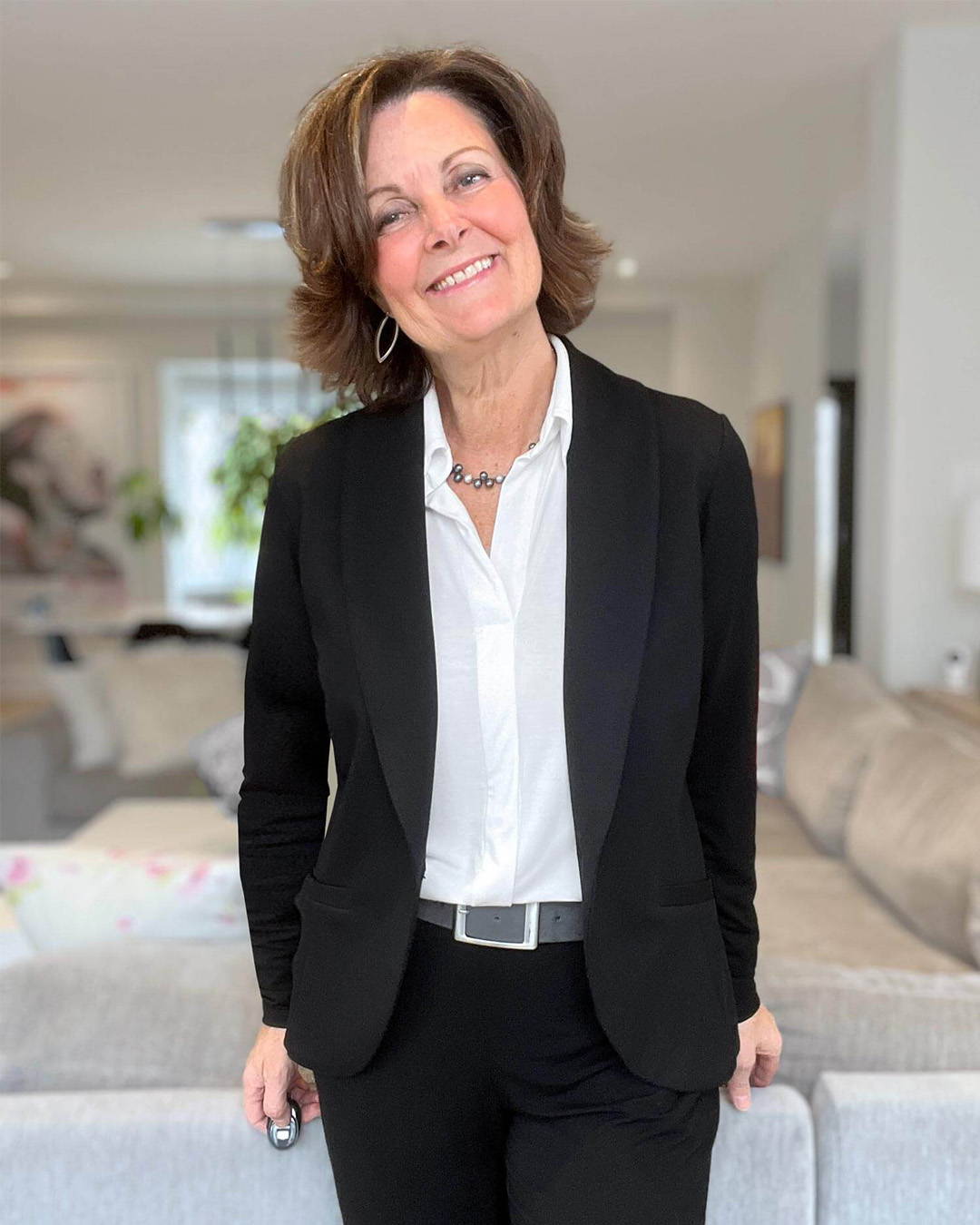 Woman standing in the living room wearing Miik's Lucia collared shirt under the Emily soft blazer in black with black pants.