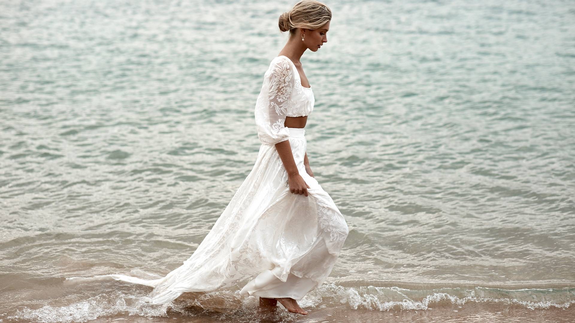 La mariée Grace Loves Lace marche dans l'océan en portant la robe de mariée Behati.