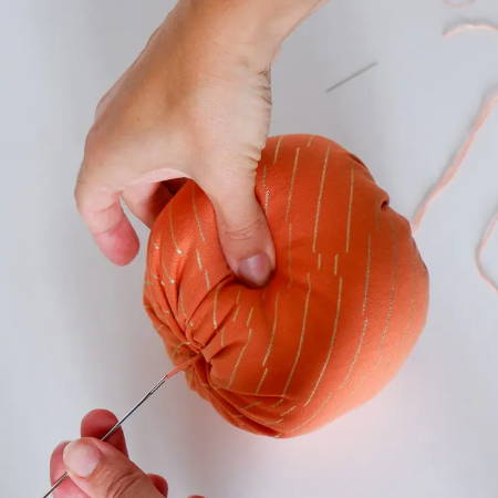 Hand pulling a big needle threaded with embroidery floss through the stuffed fabric pumpkin