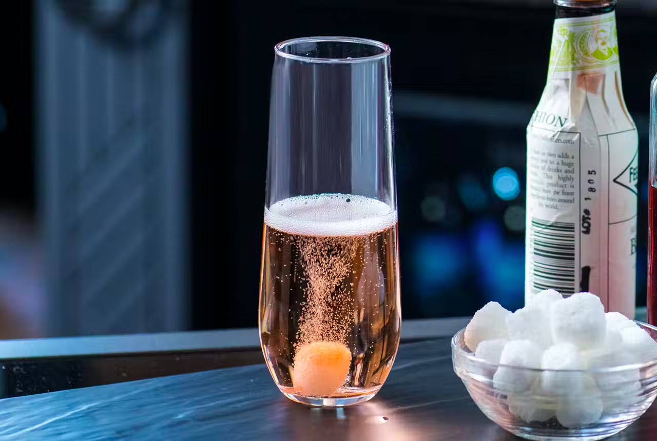 Stemless champagne glass with sparking pink cocktail and a sugar cube. Bowl of sugar cubes and a small bottle of bitters is also displayed.