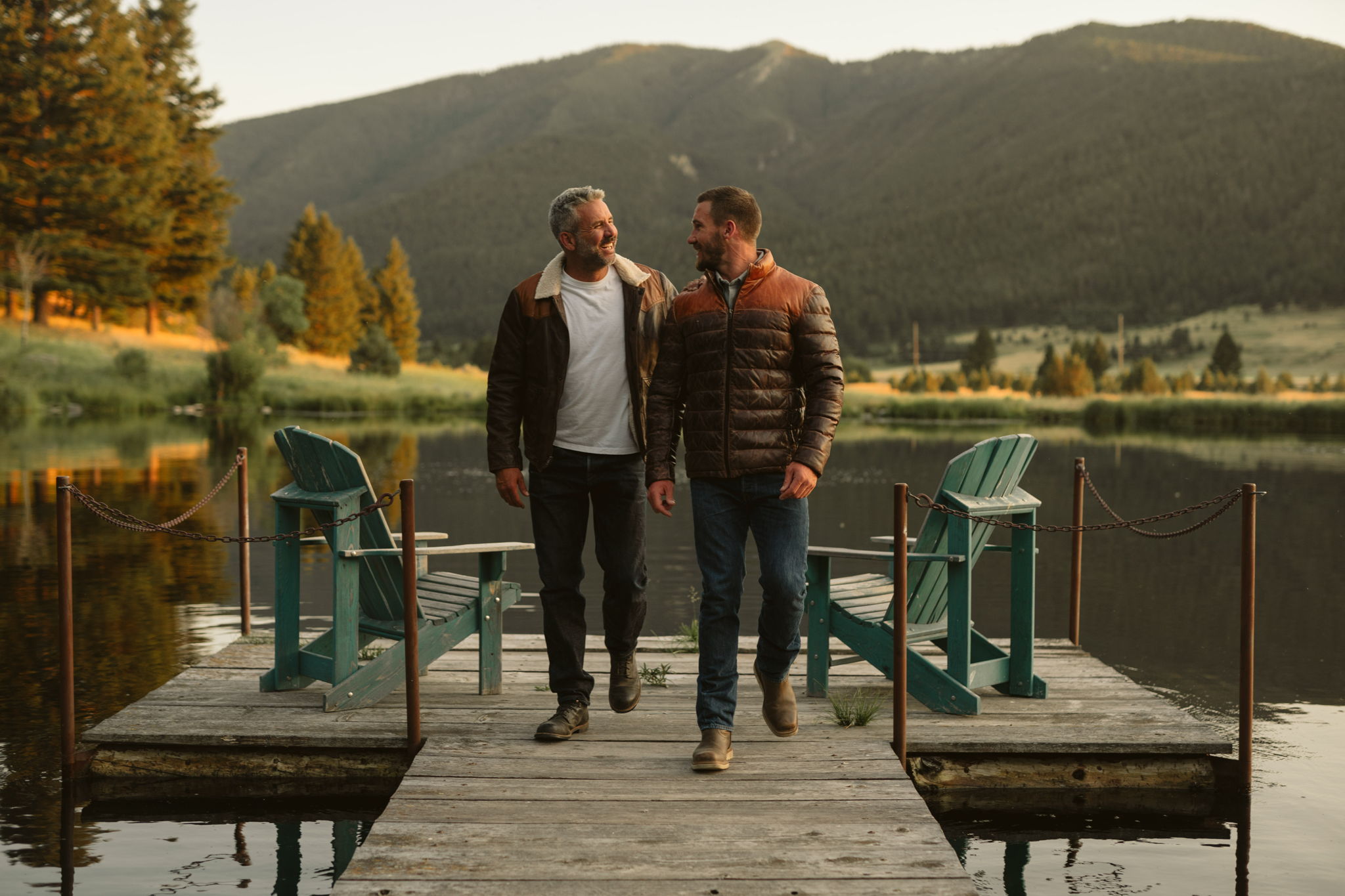 Two men in Buffalo Jackson leather jackets, one shearling-collared and one quilted down, walking on a mountain lake dock