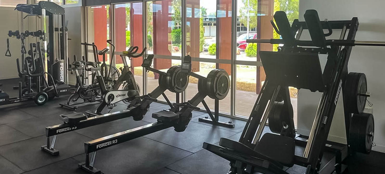 High School Gym Fit Out at St. George State. A row of indoor rowers facing the gym's large windows, emphasizing their sleek design and digital performance trackers.