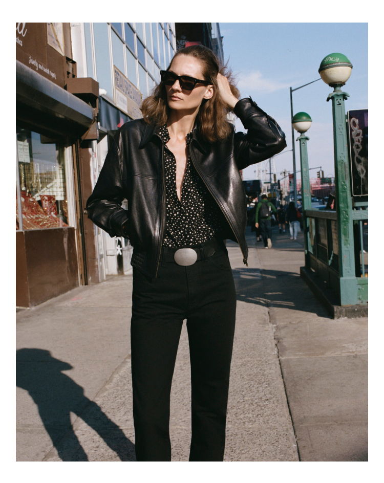 A person in a black leather jacket poses on a city street.