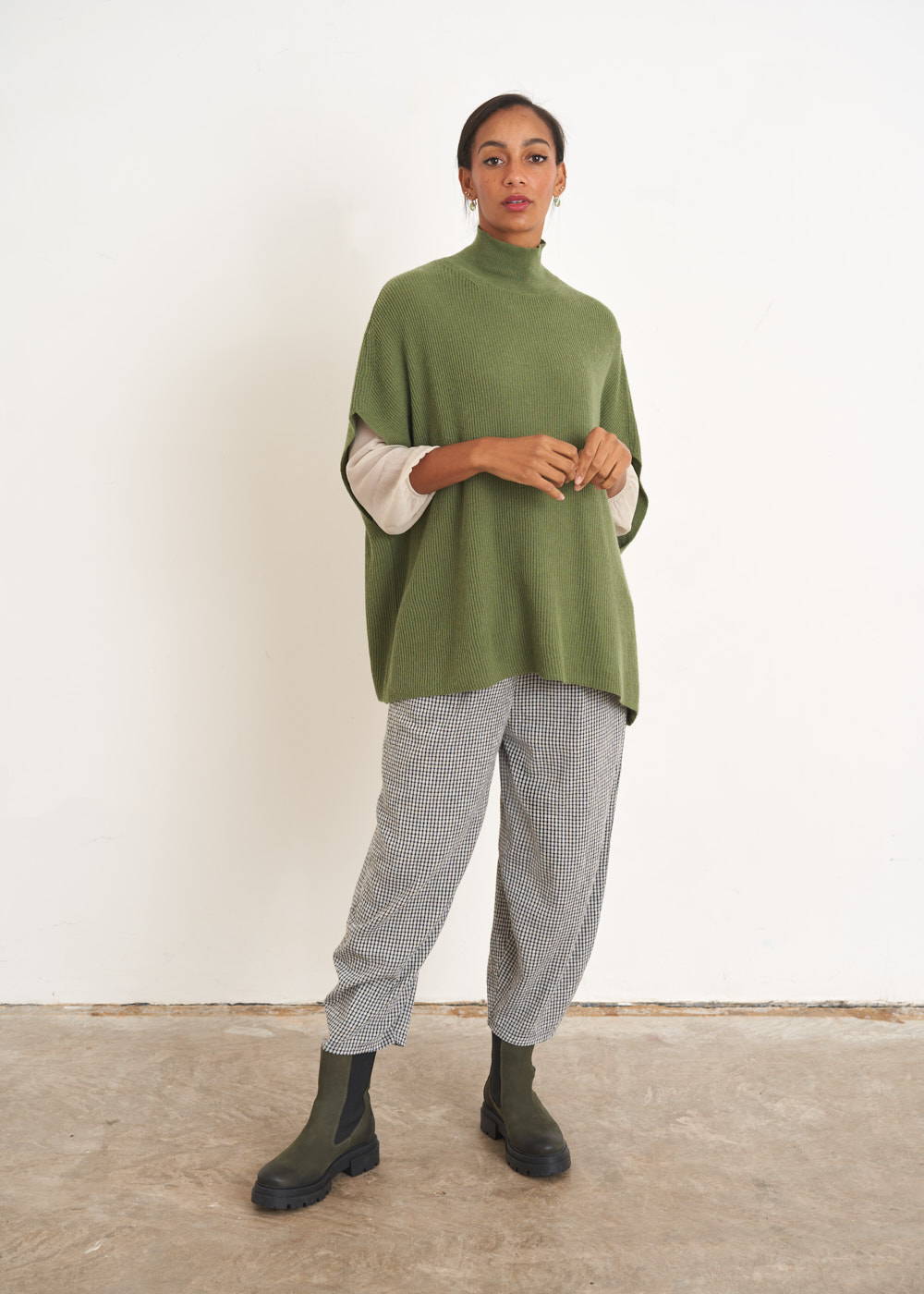 A model wearing a green knitted sleeveless sweater with a turtle neck over an off white shirt, grey checked trousers and khaki green chelsea boots