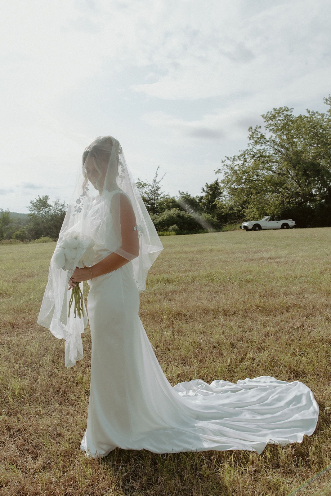 Bride in a white gown holding flowers, standing in a grassy field.