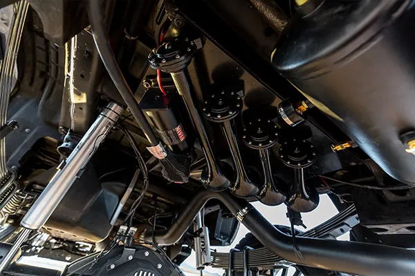 Close-up underside view of black train horn bells mounted beside suspension components on Elias's 2015 Silverado 2500HD, install photo 4