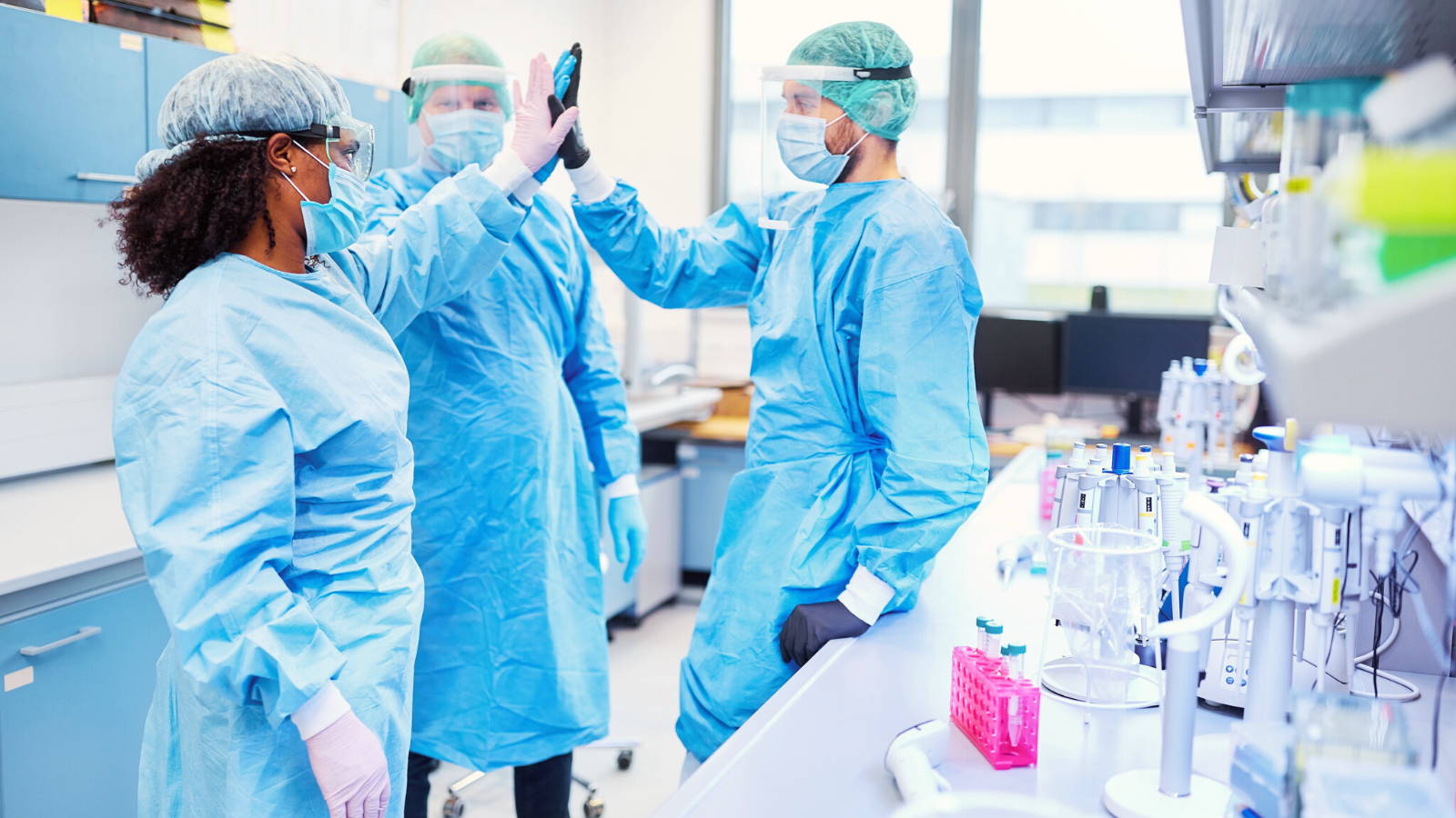 Three scientists in scrubs and masks high five each other. Photo for ethical purchasing in sciences article