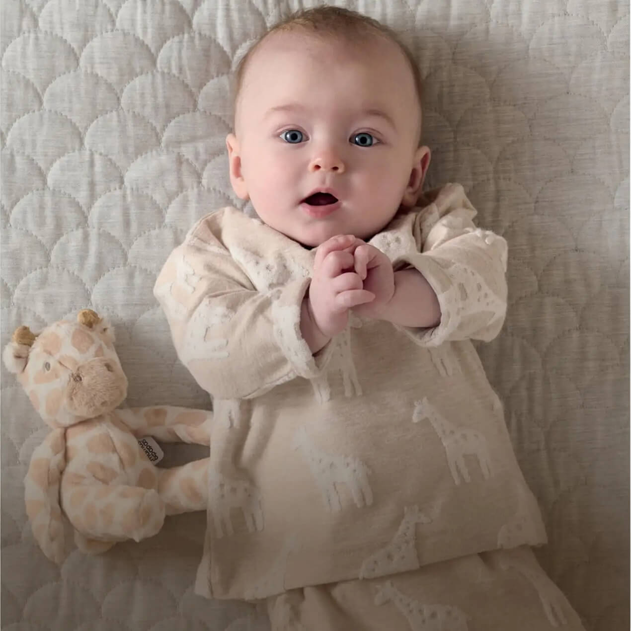 Baby in cream outfit lying on a quilt with a teddy bear.