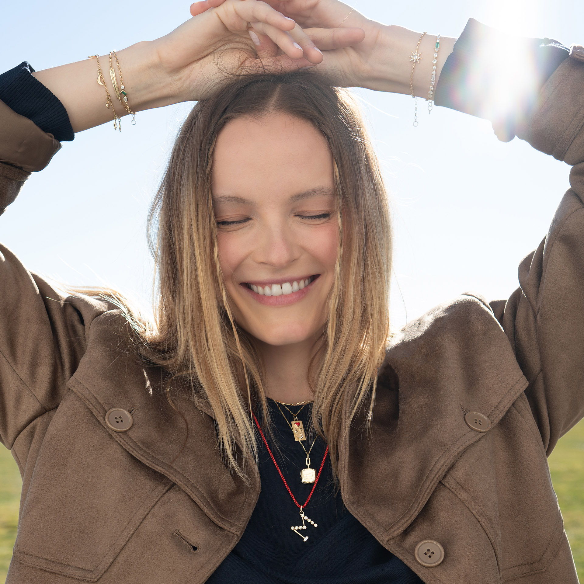 A smiling person with long hair, raising hands against a sunny background.