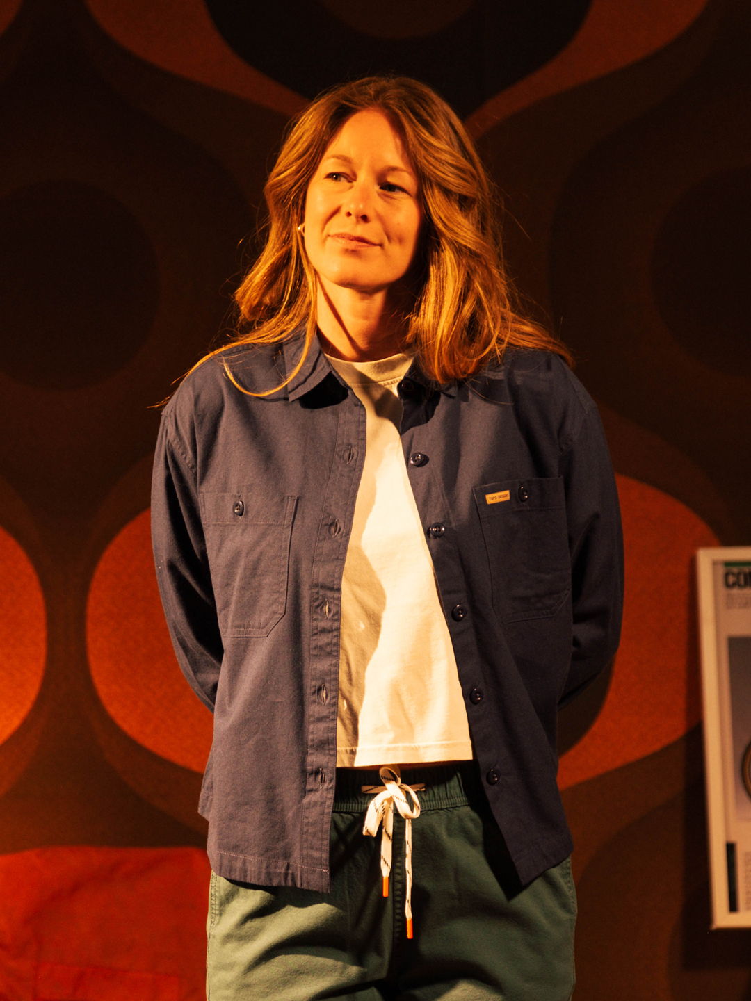 Person with long hair wearing a blue shirt and green pants, standing.