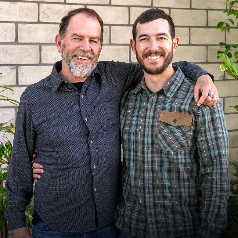 Two men smiling together in front of a brick wall.
