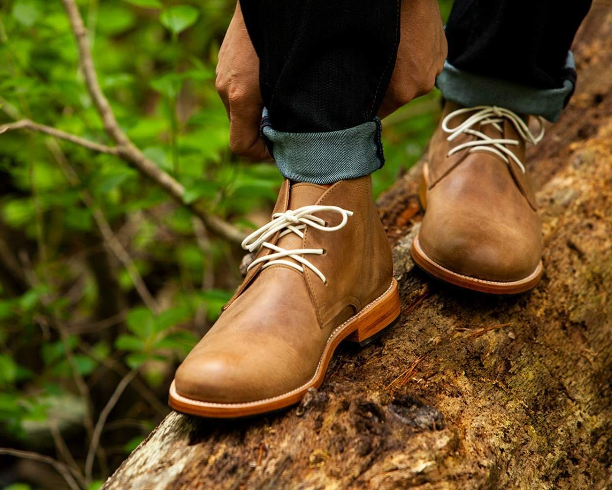 A close-up lifestyle shot of a man standing on a fallen mossy tree log in a forest, wearing light brown or desert tan leather Adelante Santiago chukka boots. The boots feature cream-colored contrast laces and a stacked leather sole. The person is adjusting the cuff of their dark denim jeans, with green woodland foliage and trees softly blurred in the background.