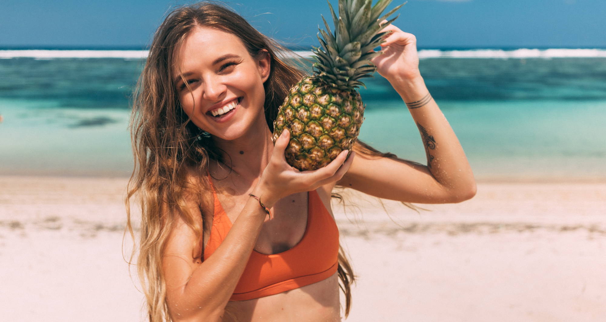 A smiling woman with long blond hair wearing an orange bra stands on a beach holding a pineapple up near her face. 