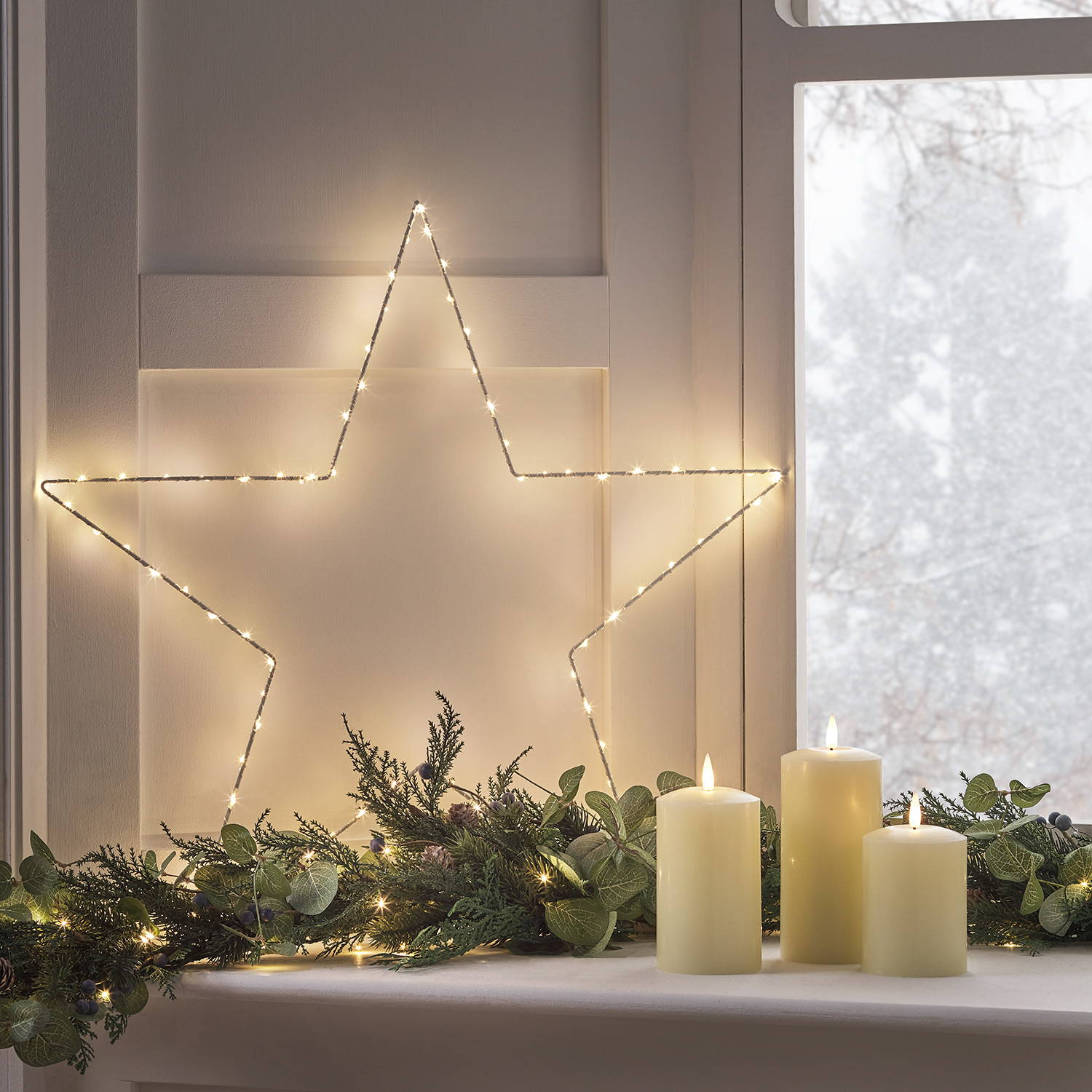 A large Osby star light with LED lit garland and candles resting on a window sill while snow falls outside