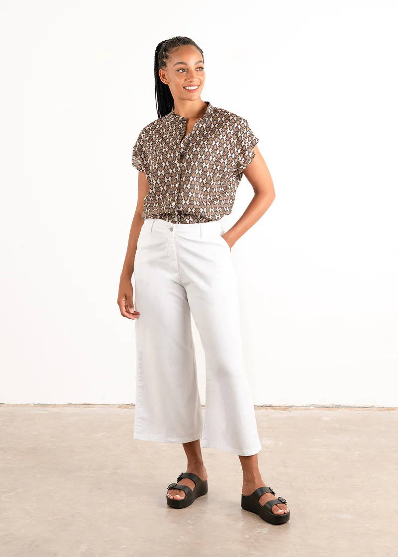 A model wearing a pair of white, cotton, wide leg 3/4 trousers with black chunky slides and a brown patterned shirt