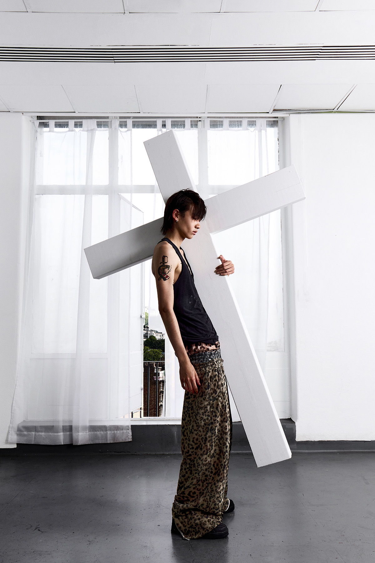 Model wearing a black tank top and leopard-print pants carrying a large white cross in a minimalist studio setting