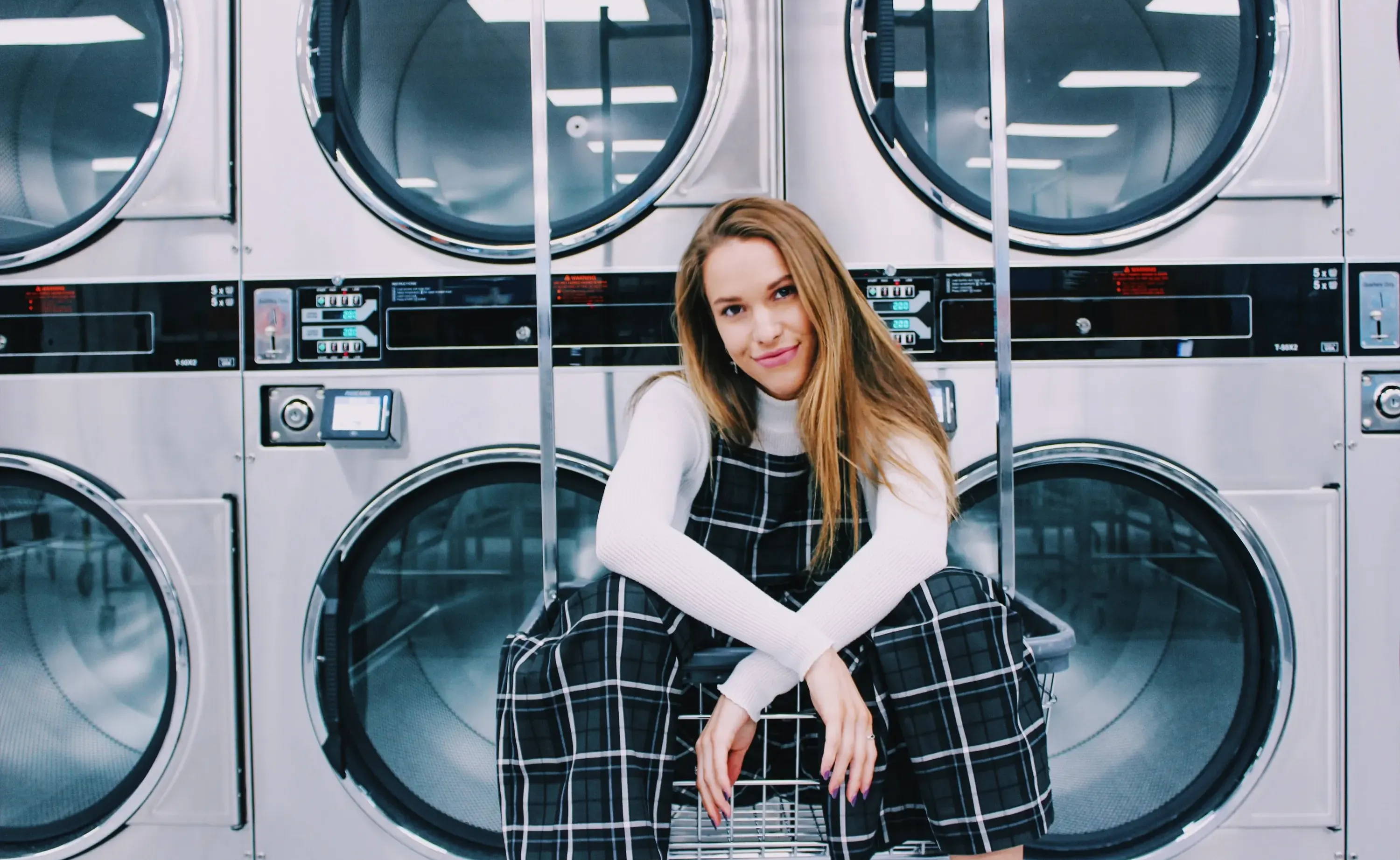 A woman in plaid overalls sits and smiles in front of laundromat washing machines.