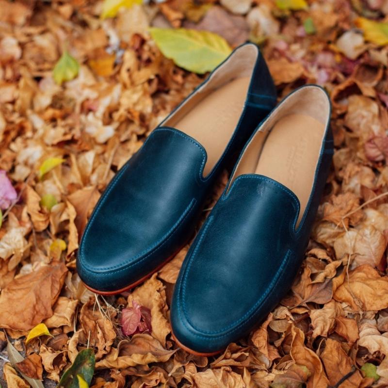 A pair of Adelante deep blue leather wide width slip-on loafers resting on a bed of dry autumn leaves.