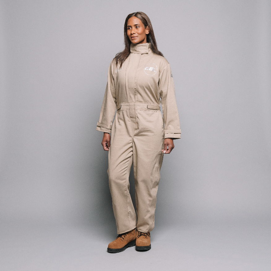 A woman in a beige jumpsuit stands against a gray background.