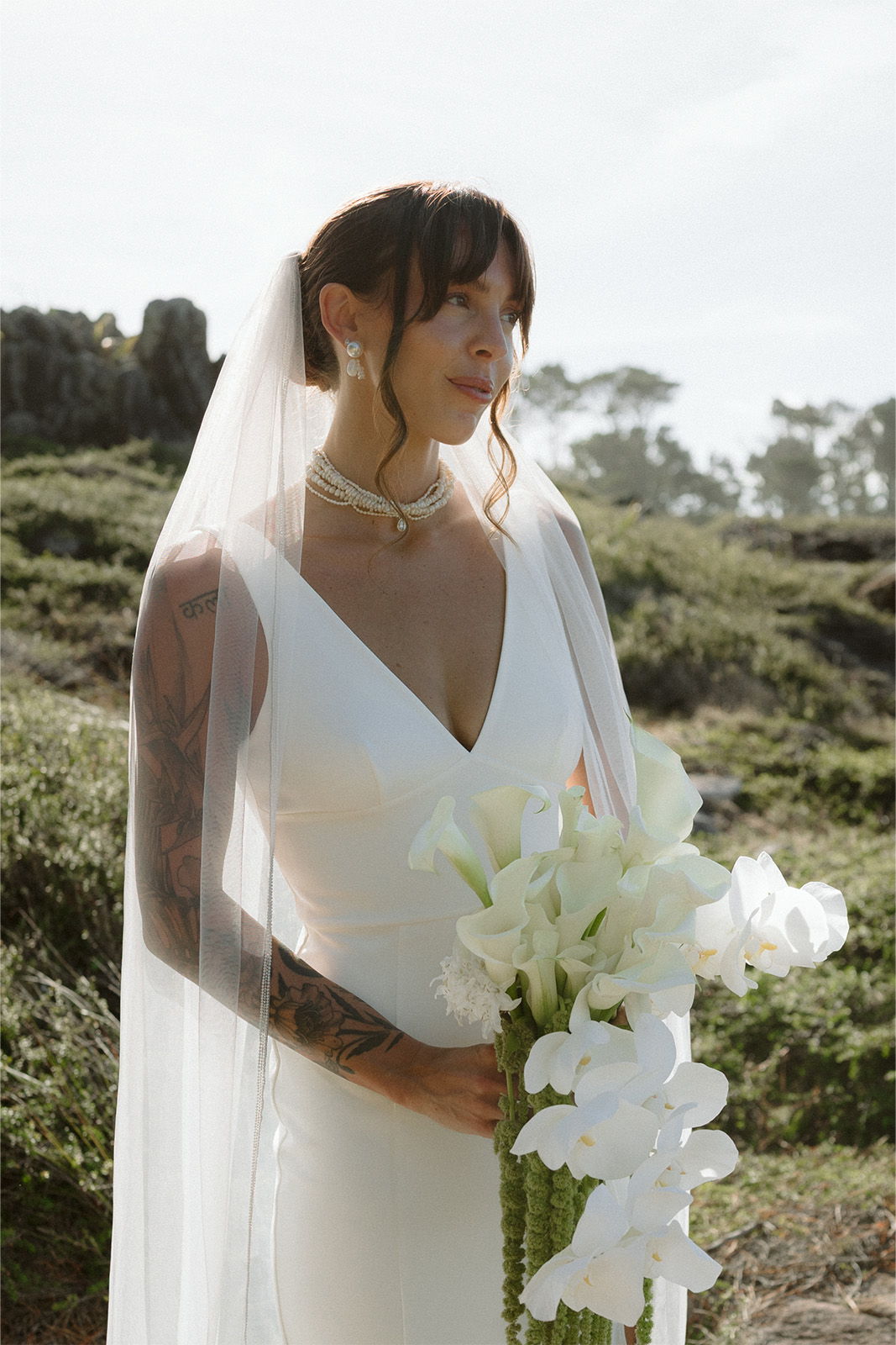 Bride in a white gown holding white flowers, with a veil outdoors.