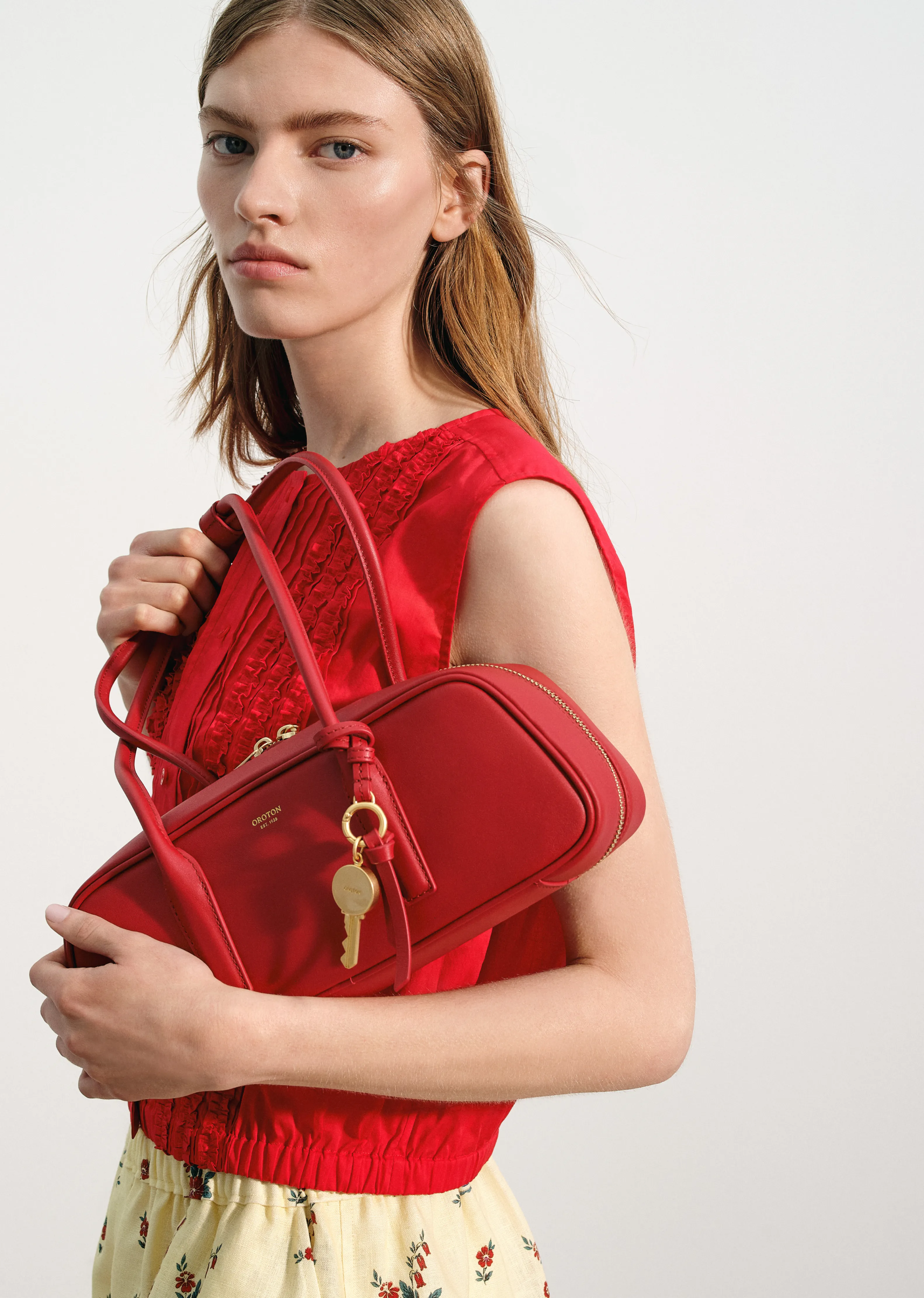 Model holding a red handbag, wearing a red top and floral skirt.