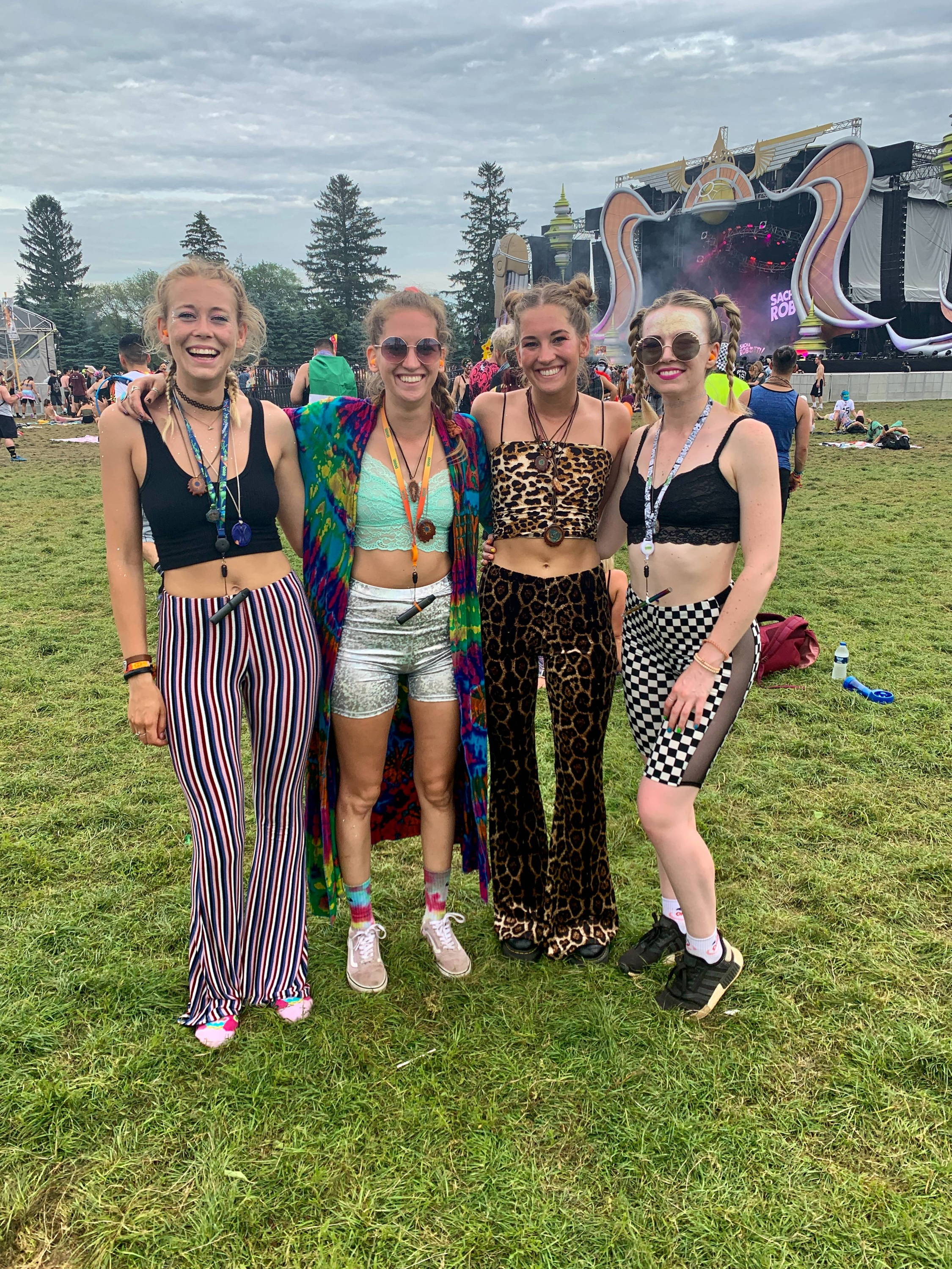 Four white girls are posing together with a stage at a music festival in the background. 3 of the girls have Ooze lanyards with vape pens around their necks.
