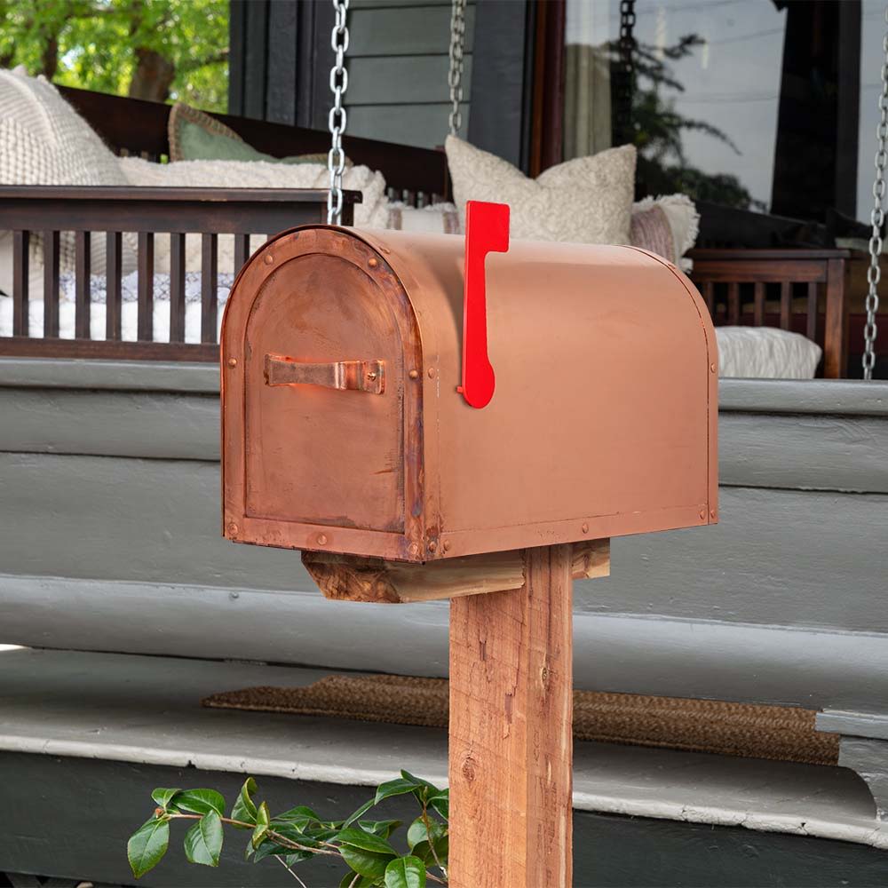 A traditional, rounded mailbox with red flag and a raw copper finish on a wood post.