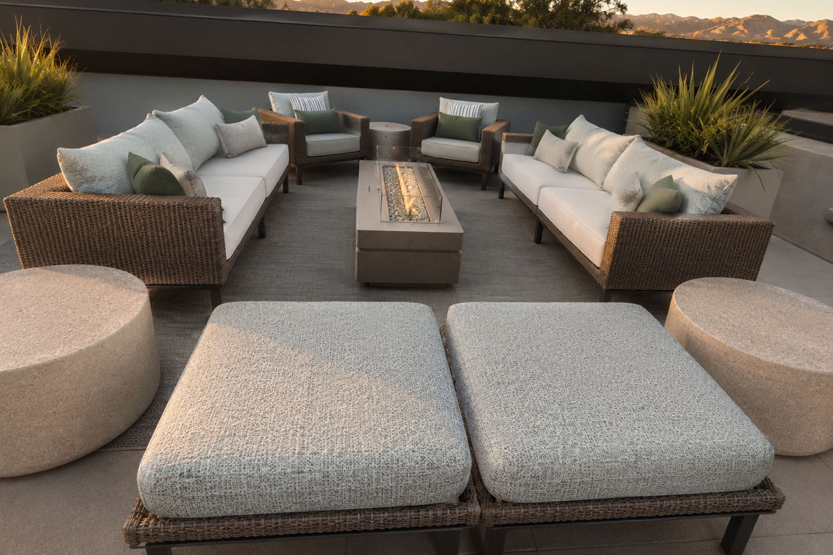 Rooftop outdoor lounge with resin wicker sofas, pale blue performance cushions, fire table, and mountain views in a warm desert setting.