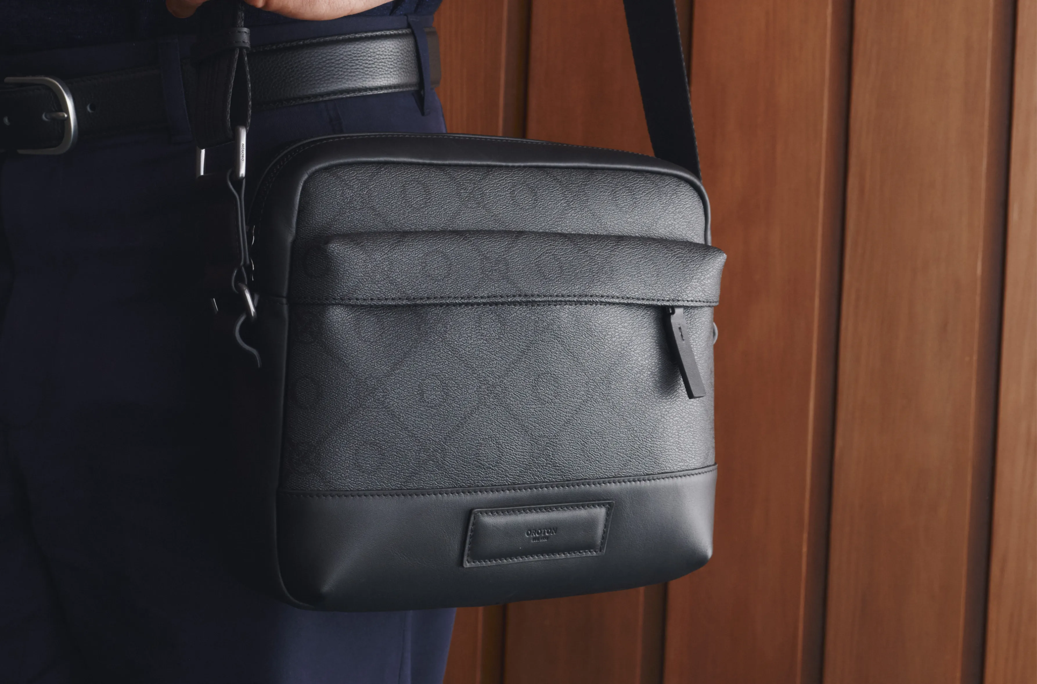 A person holding a black patterned crossbody bag against a wooden background.