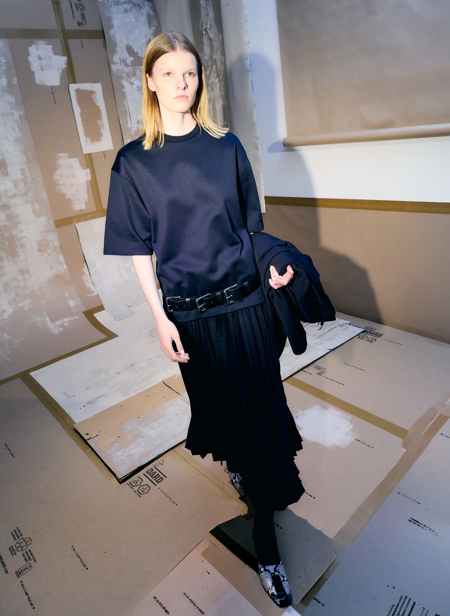 Model wearing a dark outfit poses on a runway with cardboard backdrop.