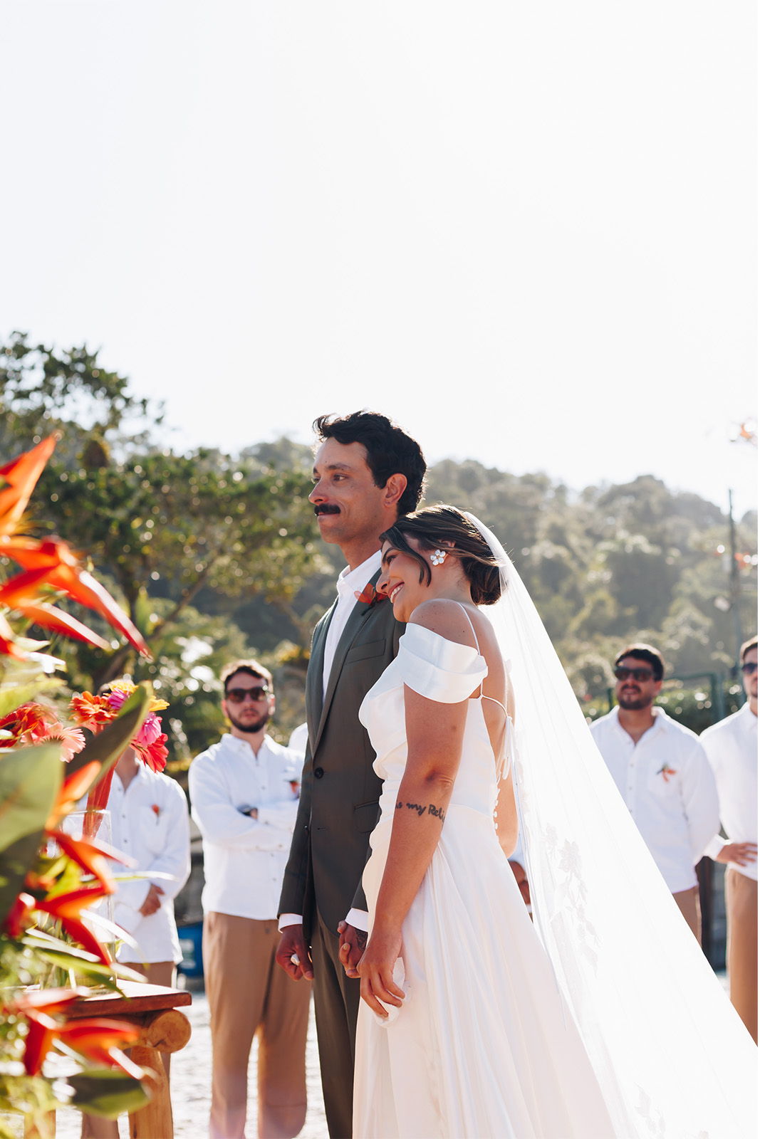 A couple in wedding attire standing together, surrounded by guests in bright attire.