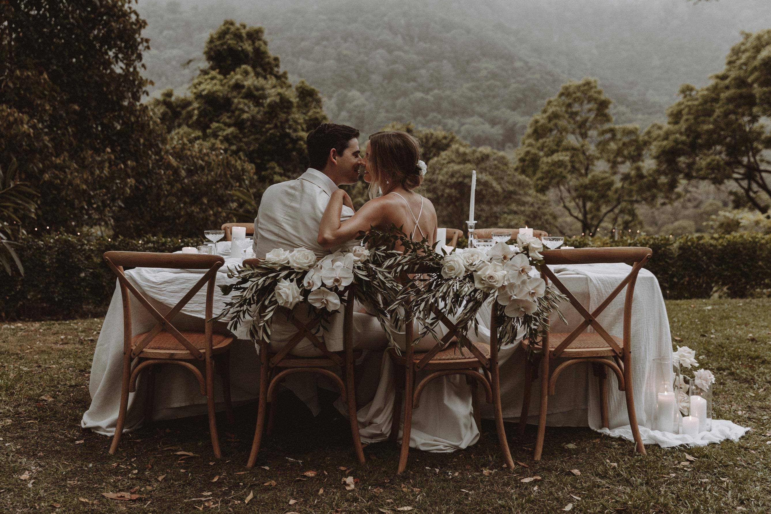 Mariés assis à leur table de mariage partageant un baiser avec des montagnes et des arbres de la forêt au loin.