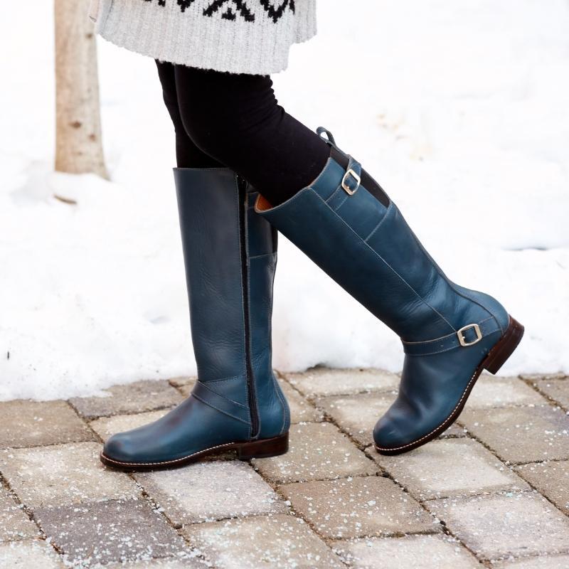 Woman wearing navy blue leather Reina wide calf equestrian boots with brass buckle details standing on a brick walkway in the snow.