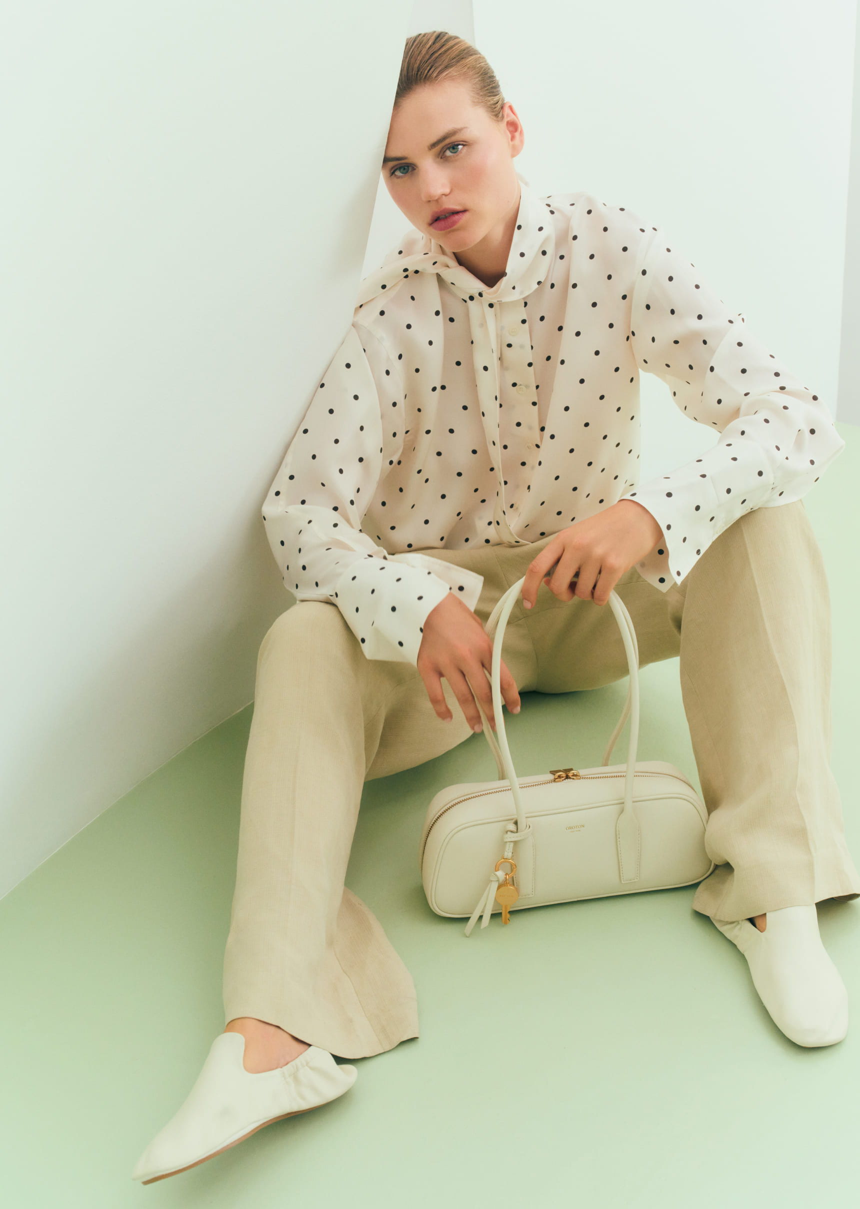 Model in stylish outfit, seated with handbag against a light-colored background.