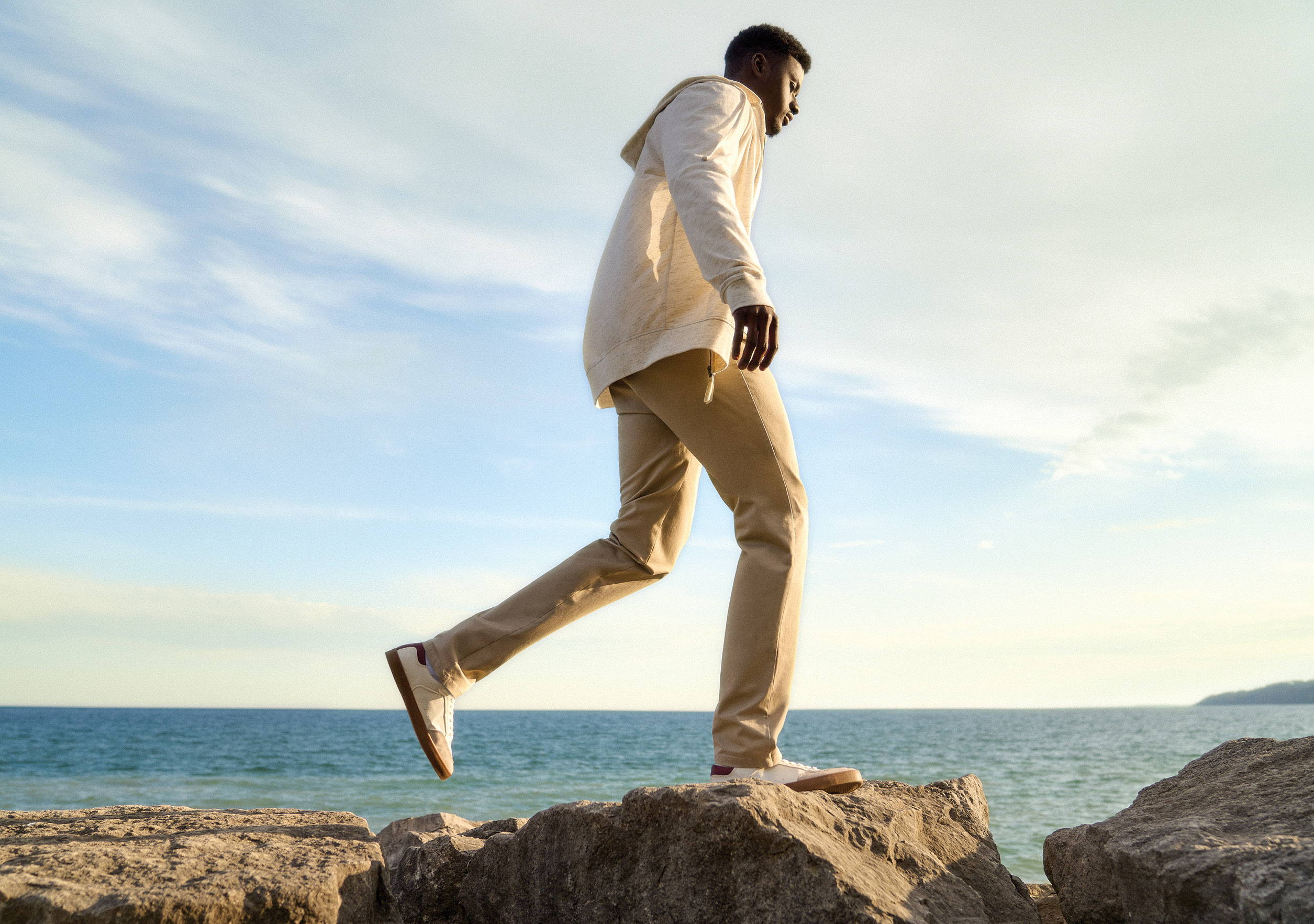 Tall man walking on rocks by the water wearing a beige sweater and khaki pants