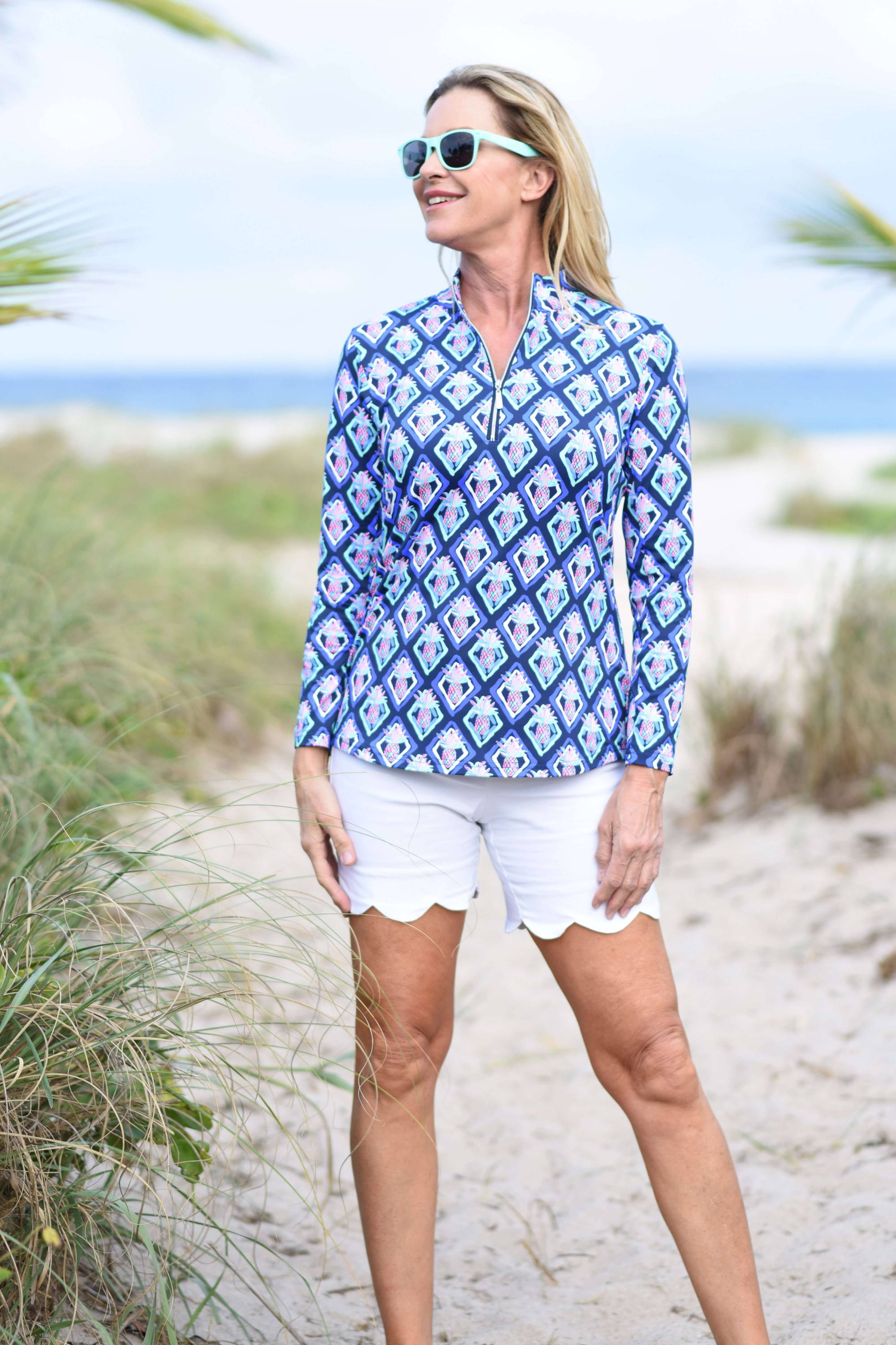 Blonde woman in sunglasses smiles and looks to the side as she stands on a beach. 