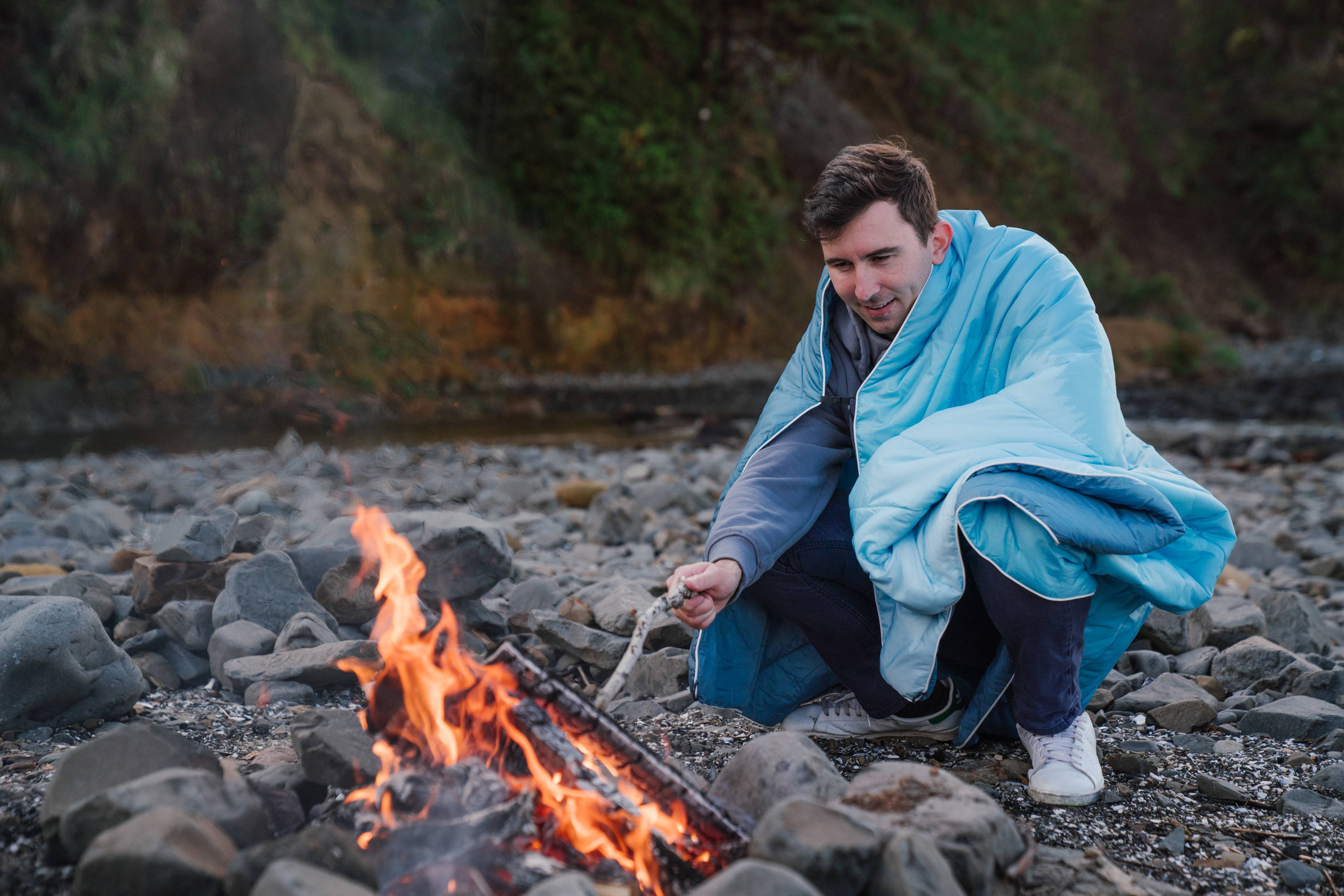 Man wearing Rumpl blanket holding stick over fire