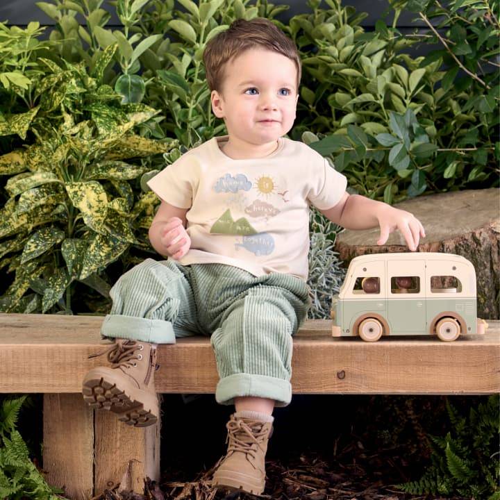 A little boy sits on a wooden bench outside, with plants behind, and is wearing a Mamas and Papas t shirt and cord trousers.