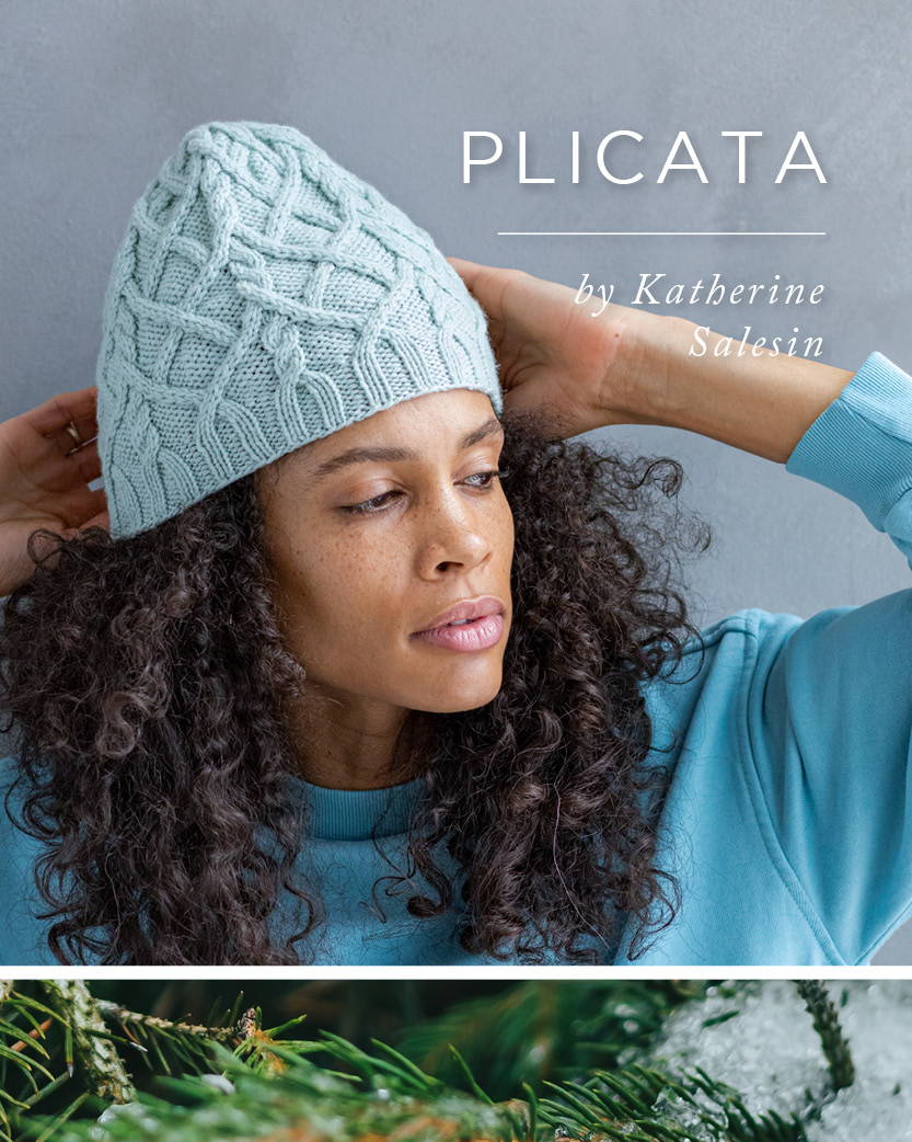 A woman with curly hair models a hand knit cabled beanie hat with the title PLICATA by Katherine Salesin