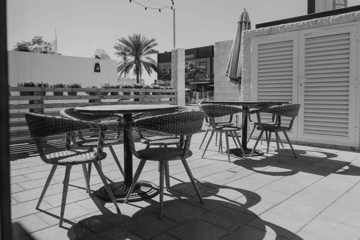 Black and white patio dining scene with woven outdoor chairs and tables in full sun, showing heat exposure on outdoor furniture in a commercial setting.