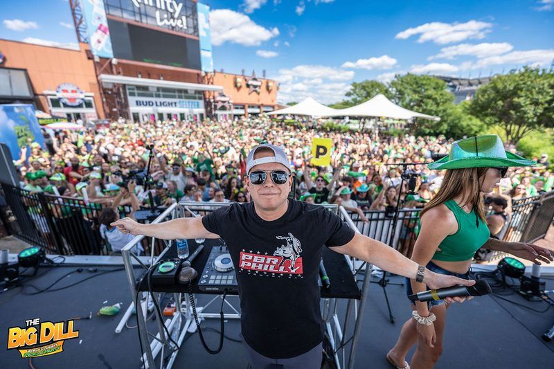 DJ in a PBR Philly shirt with arms out on the main stage in front of a sold-out crowd in green at The Big Dill® Philadelphia pickle party, returning to Philly on National Pickle Day at Stateside Live!