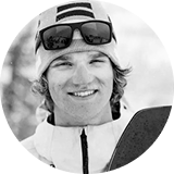 Smiling snowboarder wearing sunglasses and a hat, standing in a snowy environment.