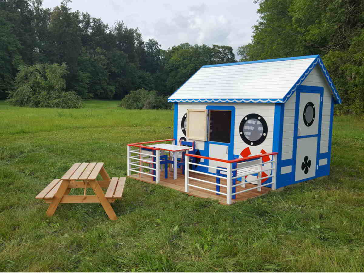 Custom playhouse like a ship in white and blue colors with railing and terrace in a grass field in front og a forest by WholeWoodPlayhouses