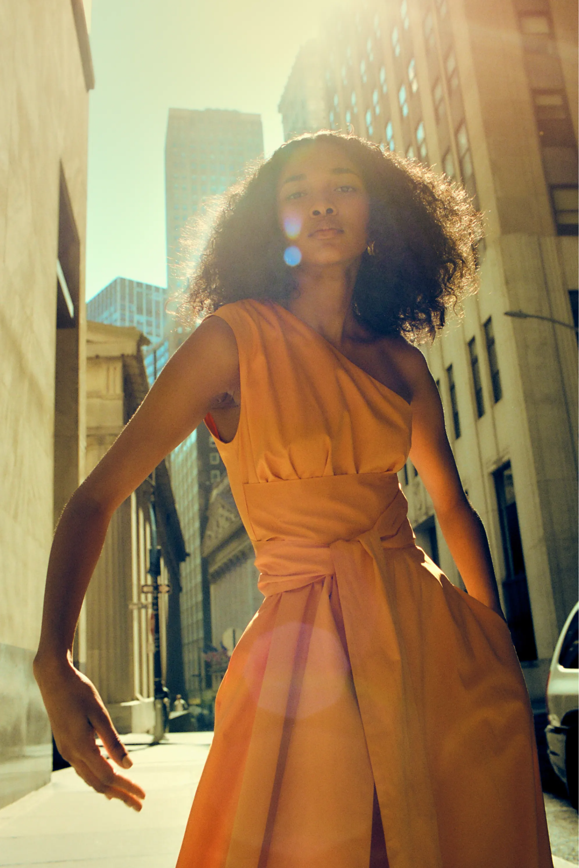 Person in an orange dress walking in a city with sunlight.