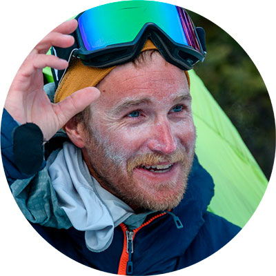 Smiling man wearing goggles, outdoors with camping gear in the background.