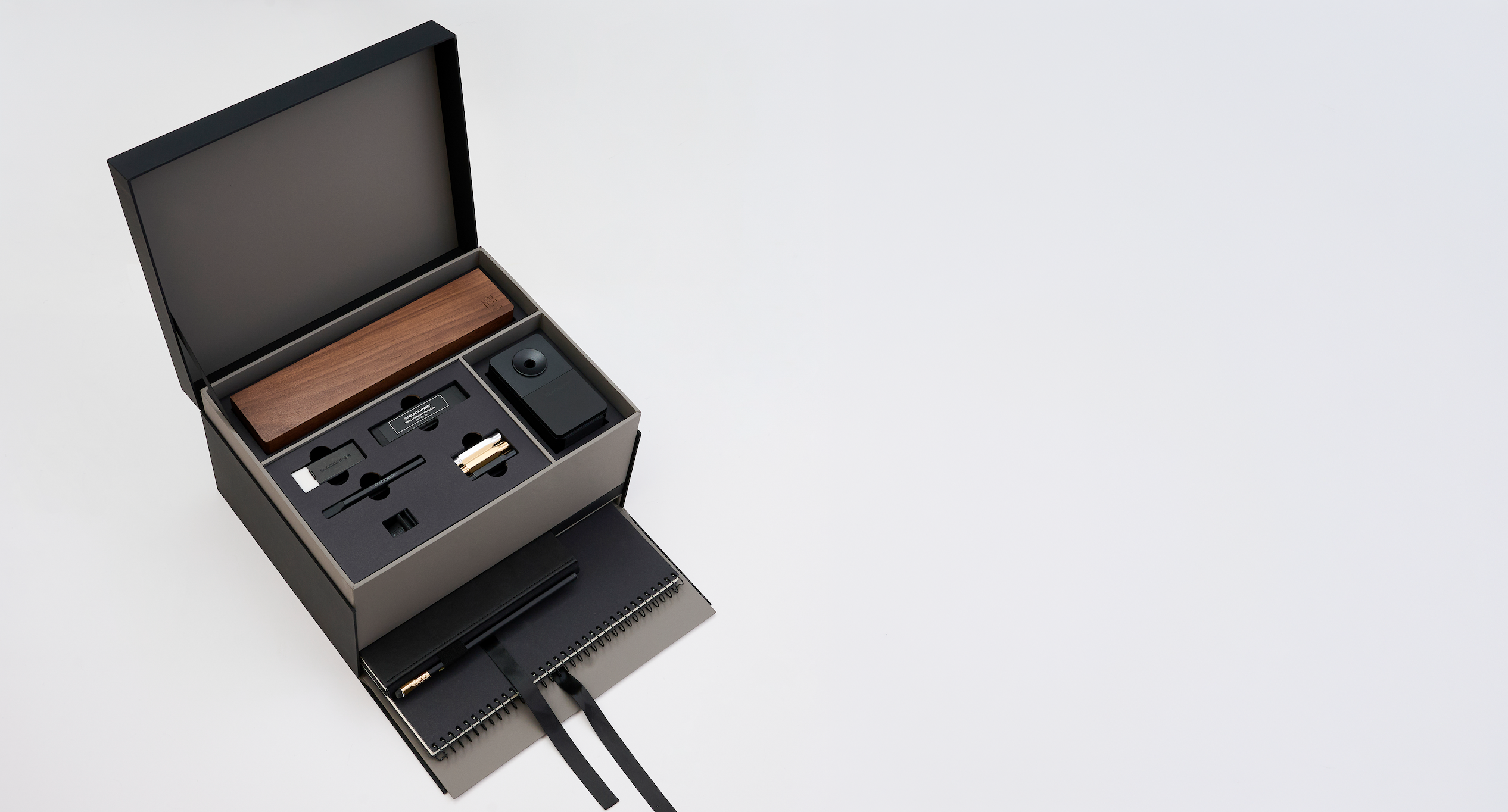 An open black gift box on a light gray background, revealing a wooden pencil box display set, blackwing handheld eraser, blackwing replacement erasers, blackwing point guards, and blackwing notebook clip on the top layer, and a large black spiral bound notebook and medium hardcover notebook with a black pencil in the holster in a lower pull-out drawer with a ribbon string.