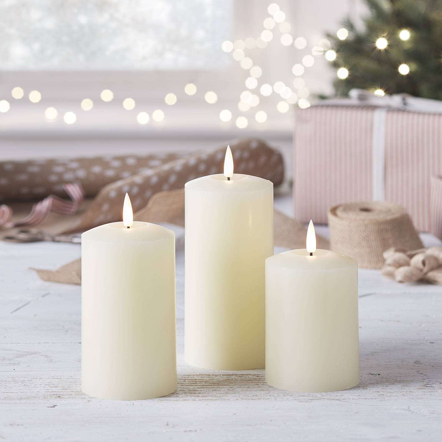 3 ivory LED pillar candles on a table with gift wrapping materials in the background.